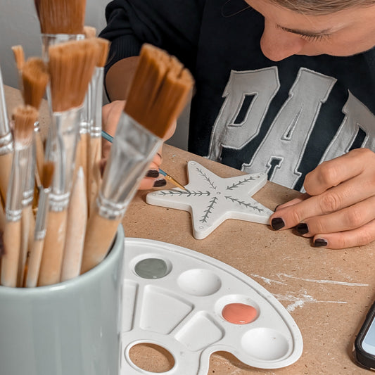Person painting a star-shaped object with brushes and a palette on a table.