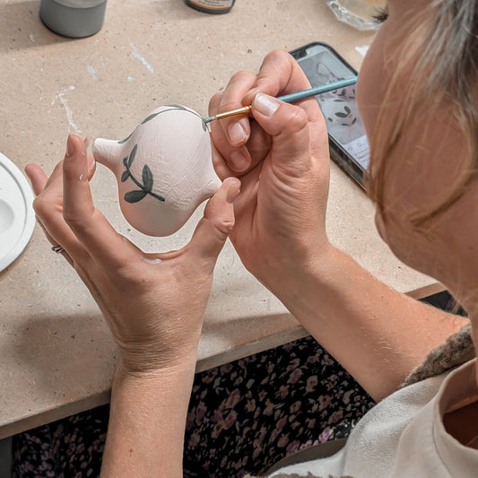 Person painting a small ceramic object with a brush on a table.